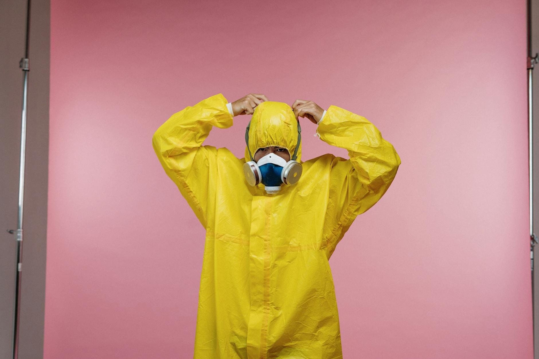 pexels-photo-3951417-3951417-1 Person wearing a yellow protective suit and respirator against a pink studio background.